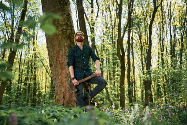 Holding an axe. Bearded man is in the forest at daytime.