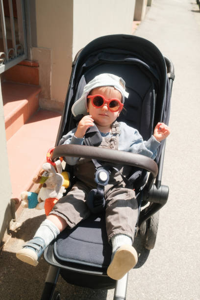 An hip baby boy is sitting in his stroller. He's wearing sunglasses and a cool hat.