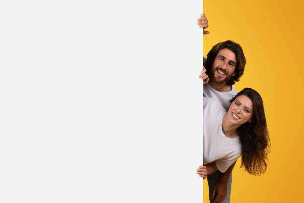 istock Joyous couple playfully peeking around a blank white space for copy or advertisement 2153049454