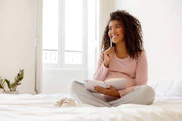 istock Happy Black Pregnant Woman Writing Checklist In Notepad While Sitting On Bed 2153050326