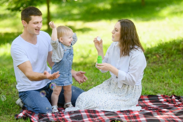 istock Dad and child are having fun, catching soap bubbles that mom blows while sitting on blanket in park on picnic. Summer time concept, outdoor recreation, weekend. Family day 2153054015