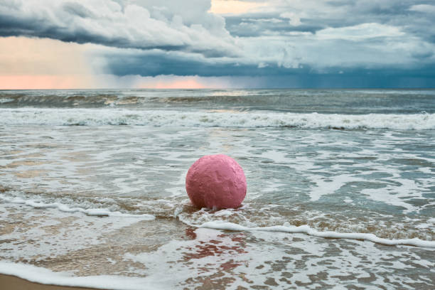 Large pink sphere lapped by waves against seashore against horizon and storm clouds, pink ball symbol of serenity contrasts against brewing turbulence in sky and relentless energy of sea