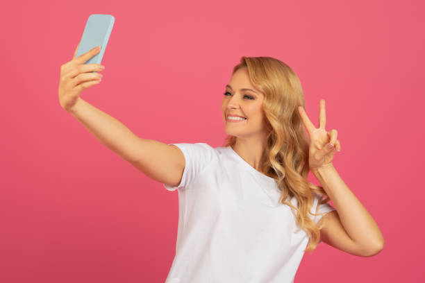 istock Blonde lady posing with smartphone for selfie showcasing victory, studio 2153098363