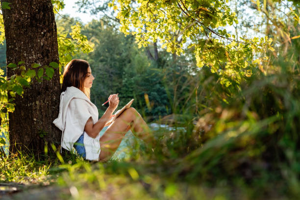 istock Young attractive girl writing and drawing in notebook sitting under the tree on nature in sunlight. Closeness to nature  inspires creativity. Urban escapism. 2153113030
