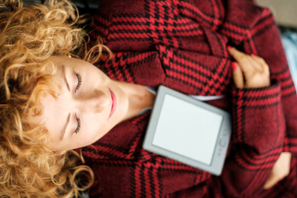 Elevated portrait of relaxed woman with eyes closed and there is an e-reader on her. Curly red hair and red plaid coat, her arms are crossed around of the e-book.