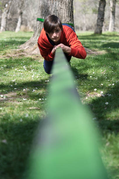 Concentrated young boy looking at camera while lying on slackline tied to trunk and crawling with help of hands in green forest in daylight