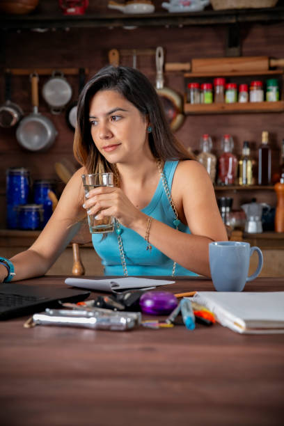 istock Happy Hispanic freelancer young woman sitting on a table and using laptop computer with a glass of water at home office kitchen 2153154730