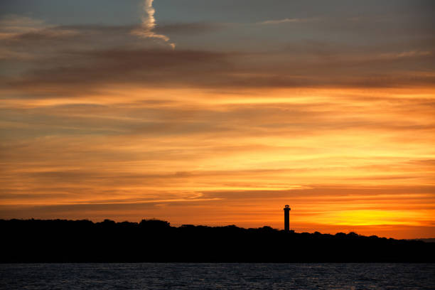 Silhouette of a lighthouse standing on a sloping coastal hill under an orange sunset