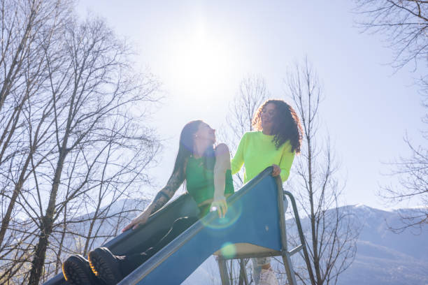 Two young women share laughter and joy as they enjoy a sunny day by the lake, sliding down a vibrant slide in the refreshing outdoors.