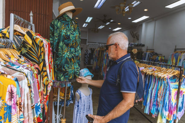 Man Browsing Tropical Shirts at Clothing Market A man inspects a tropical shirt on a mannequin at an outdoor market stall. Surrounded by vibrant garments, he enjoys a casual and detailed shopping experience. colorful check shirts on sale at a clothing store stock pictures, royalty-free photos & images