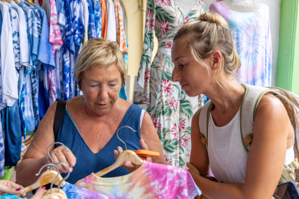 Women Shopping for Tie-Dye Shirts at Market Stall Two women browse and examine colorful tie-dye shirts at an outdoor market stall. They discuss the garments, enjoying a casual and engaging shopping experience. colorful check shirts on sale at a clothing store stock pictures, royalty-free photos & images