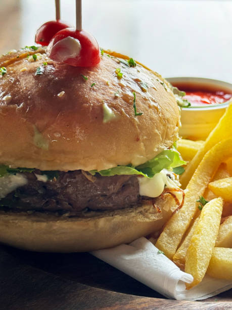 Close-up image of wooden skewered halved hamburger in seedless burger bun on wooden plate served with pile of crispy French fries, ramekin of tomato ketchup, paper napkin, focus on foreground
