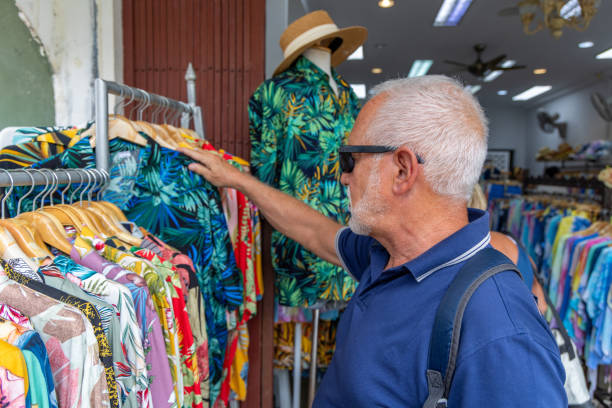 Senior Man Browsing Colorful Hawaiian Shirts at Shop Elderly man with sunglasses and white hair looking through vibrant Hawaiian shirts at an outdoor store. Mannequin in tropical shirt and straw hat in the background. colorful check shirts on sale at a clothing store stock pictures, royalty-free photos & images