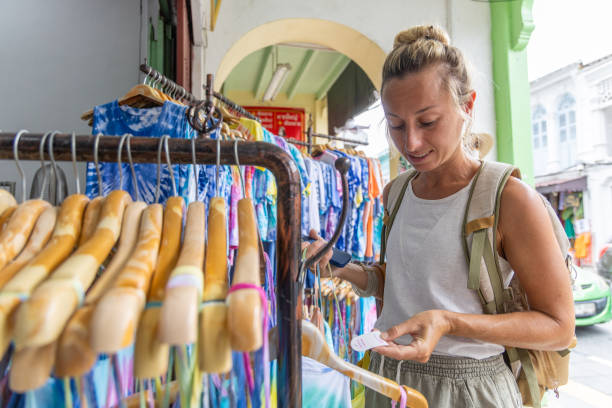 Woman Checking Price Tag at Outdoor Clothing Market Woman with a backpack examining the price tag of a tie-dye dress at an outdoor market. Racks filled with colorful tie-dye apparel in the background. colorful check shirts on sale at a clothing store stock pictures, royalty-free photos & images