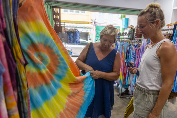 Women Shopping for Vibrant Tie-Dye Fabrics Two women examining a colorful tie-dye fabric in a boutique, enjoying their shopping experience. colorful check shirts on sale at a clothing store stock pictures, royalty-free photos & images