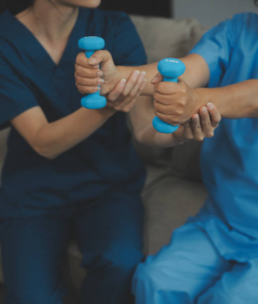 istock Old woman training with physiotherapist using dumbbells at home. Therapist assisting senior woman with exercises in nursing home. Elderly patient using dumbbells with outstretched arms. 2153205404