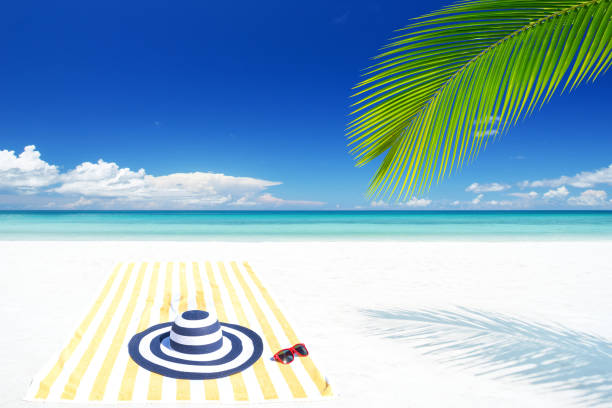 A relaxing beach setup with sunglasses, citrus fruits, and a blue cocktail resting on a striped towel. The turquoise sea and clear sky create a serene spa-like vacation vibe.