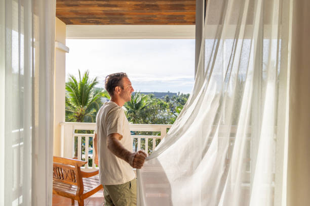 istock Man Opening Curtains to Bright Tropical Morning 2153237237