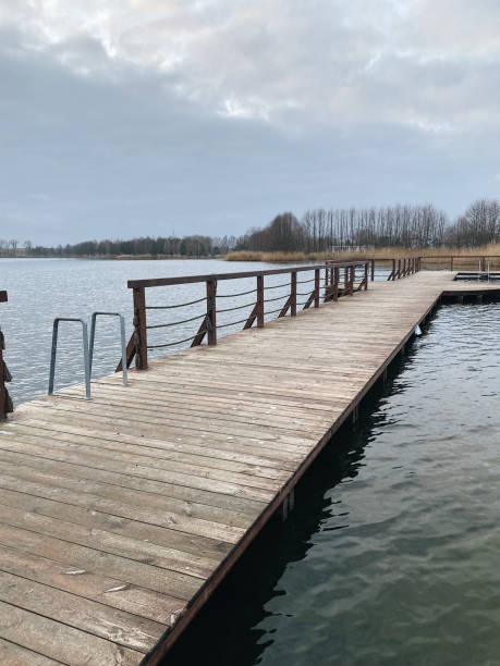 istock Wooden pier extending into a calm lake under a cloudy sky, surrounded by bare trees and reeds, creating a peaceful, serene landscape 2153239043