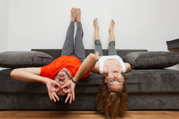 A young man and woman enjoy a fun moment hanging upside down on their home sofa, displaying playfulness and joy in a cozy setting.