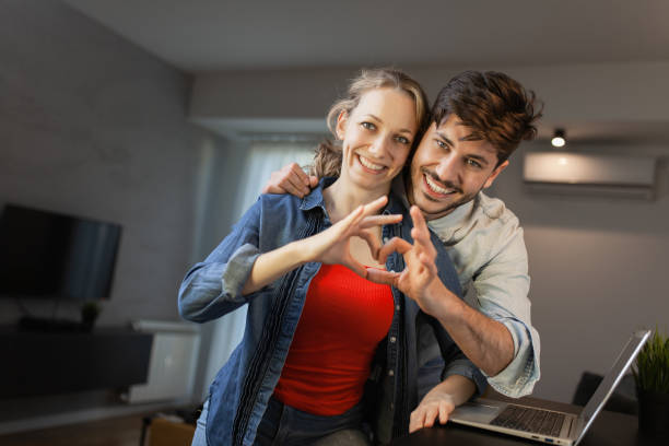 istock A joyful young couple forms a heart shape with their hands, smiling warmly in their modern living room with a laptop in the background. 2153244815