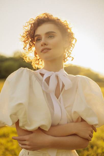This evocative image depicts a young woman with curly red hair walking through a sunlit meadow during late afternoon. Her vintage-style white dress, with puffed sleeves and a tie at the neck, adds a timeless elegance to the scene. She appears absorbed in the moment, gently touching the wildflowers as she walks by. The warm, natural lighting and the open field background enhance the pastoral and peaceful ambiance, evoking a sense of freedom and connection to nature.