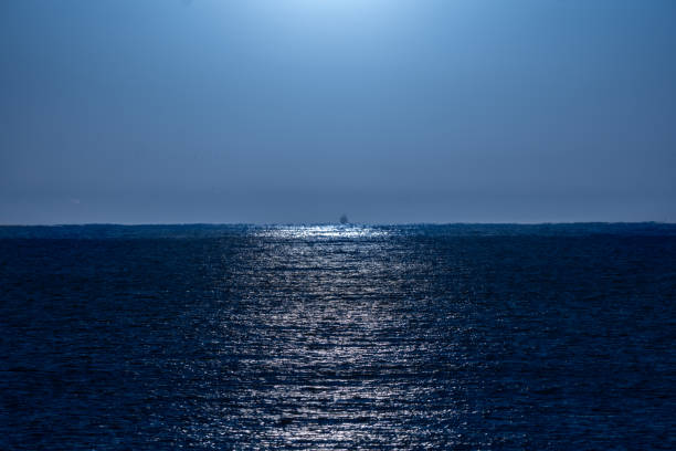 Clear, bluish night sky illuminated by moonlight with its rays of light beautifully reflected in the water of the Mediterranean Sea and a backlit fishing boat silhouette on the horizon line.