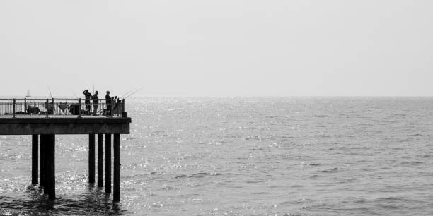 A group of fishermen standing in the distance and in silhouette on the end of a pier on the East Coast of England on a cloudless summer’s day