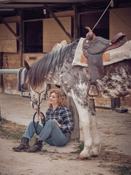 A stylish cowgirl poses in a rustic barn doorway, wearing a patterned jacket, blue jeans, belt and green boots. The relaxed stance conveys western charm, independence, and country vibes.