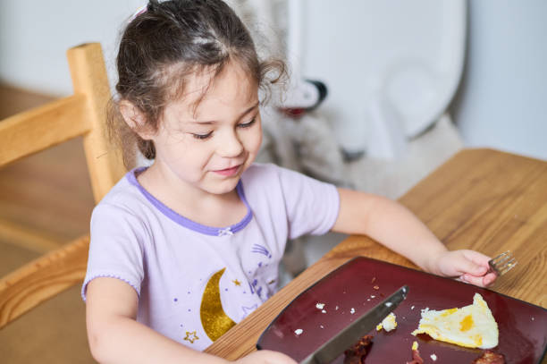istock Young pretty girl eating eggs and bacon with knife and fork for the first time 2153302536
