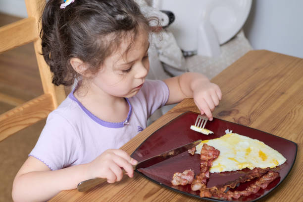 istock Young pretty girl eating eggs and bacon with knife and fork for the first time 2153302537
