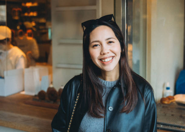 This photo captures a solo traveler, an Asian woman smiling brightly in a cafe. Her cat-ear sunglasses add fun to her journey. While others are blurred, she stands out, enjoying her moment of travel solitude. esta foto captura a una viajera sola, una mujer asiática sonriendo alegremente en un café. sus gafas de sol con orejas de gato añaden diversión a su viaje. mientras que otros se difuminan, ella se destaca, disfrutando de su momento de soledad viajera.