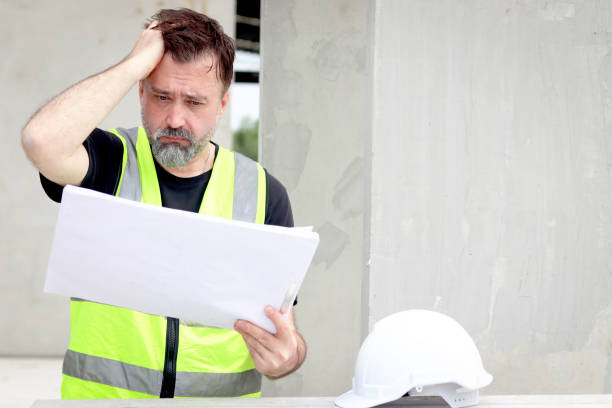istock Panic attacks senior worker engineer foreman with safety vest and helmet put hand on head while staring at blueprint at construction site. Stressed elderly architect works at building site workplace. 2153350167