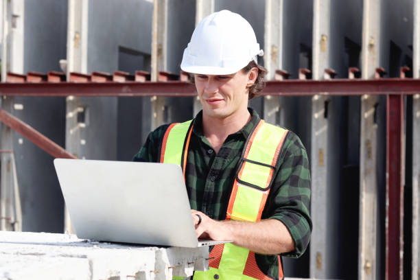 istock Happy worker engineer with safety vest and helmet typing on laptop computer while inspecting working area at construction building site. Young architect works at building site workplace. 2153350449