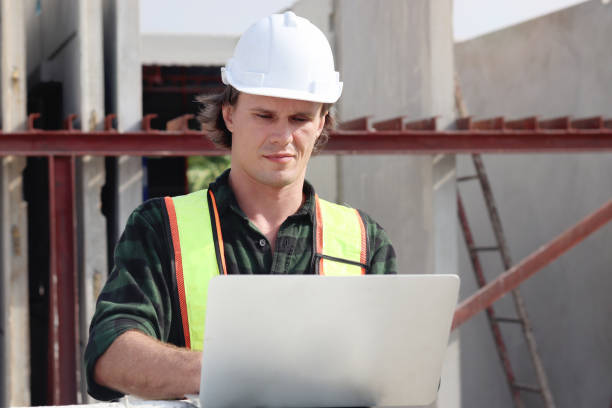 istock Happy worker engineer with safety vest and helmet typing on laptop computer while inspecting working area at construction building site. Young architect works at building site workplace. 2153350458