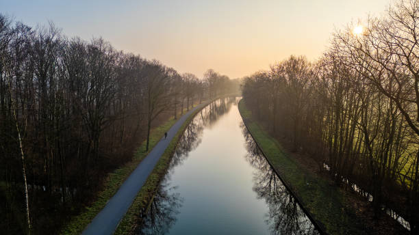 The gentle embrace of dawn illuminates a still canal, flanked by the quietude of bare winter trees. Tranquil Dawn Over Canal Flanked by Trees in Aerial Perspective. High quality photo