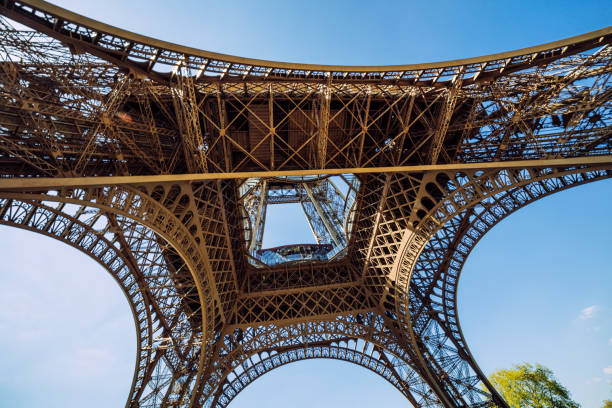 View from underneath the Eiffel Tower with sunlight shining through, showcasing its intricate ironwork design on a clear day in Paris, France.