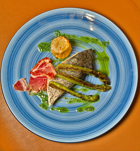 A beautifully plated gourmet dish featuring a piece of fish, grilled asparagus, prosciutto, and a corn segment, served on a striking blue plate. The vibrant colors and elegant presentation make this dish stand out, perfect for culinary blogs, restaurant advertisements, and food industry promotions. The setting includes a neatly arranged table, providing ample copy space for customized messaging.