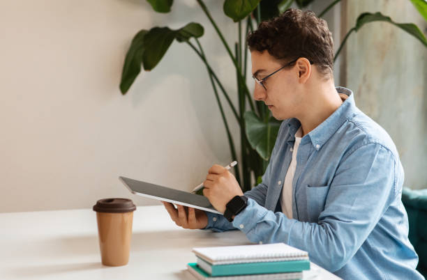 istock Focused young man with curly hair and glasses using a digital tablet and stylus to work on a project 2153361901