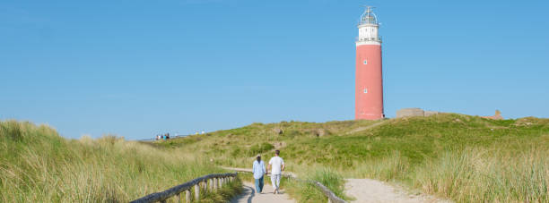 istock Two people walk down a path next to a towering lighthouse in Texel, Netherlands, with waves crashing against the shore in the background 2153365023