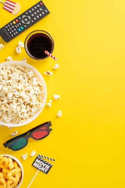 istock Enjoy movie debut at home using TV app. Top view vertical shot showcases scrumptious popcorn, snacks, soda, 3D glasses, remote control, movie-themed decor on sticks. Yellow background for adverts 2153365345