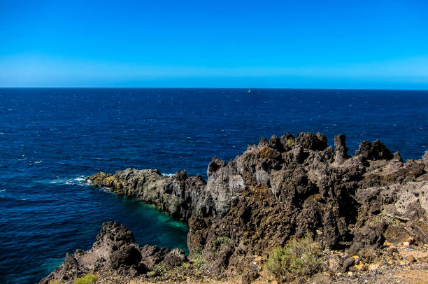 A rocky shoreline with a blue ocean in the background. The rocks are jagged and the water is calm