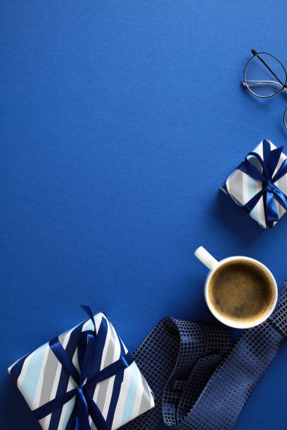 istock Stylish flat lay composition featuring striped gift boxes, glasses, coffee cup in a white mug, elegant tie on a vibrant blue background. Ideal for Fathers Day articles, promotional materials 2153367249
