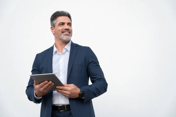 Middle age mature businessman using pc tablet for business work application. Smiling handsome older man in formal suit holding touchscreen device looking aside. Isolated white background, copy space