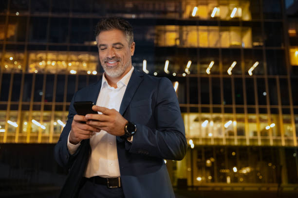 istock Smiling mature hispanic senior business man using app, holding smartphone cellphone at office centre at night. Indian or latin middle age male entrepreneur businessman working on mobile cell phone. 2153377141