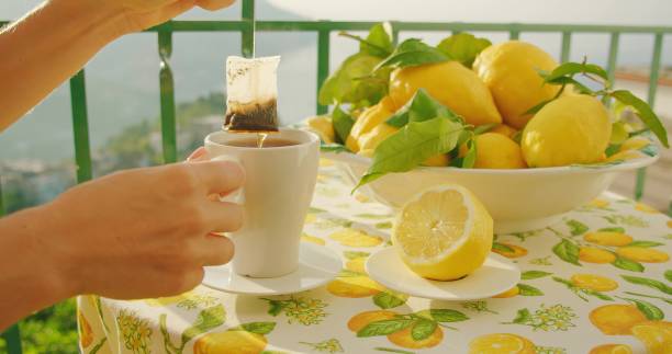 istock Tea bag steeps in a white mug on a table with lemons, mountains beyond. The act of dipping reflects a serene start to the day. Close-up of female hands. 2153377267