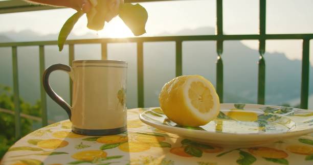 Morning light illuminates a lemon on a citrus-themed table. Hand squeeses citrus juice against coastal landscape with sea view.
