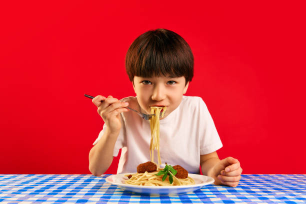 Delightful mealtime. Little boy, kid sitting and table with checkered tablecloth and eating delicious spaghetti with meatballs against red background
