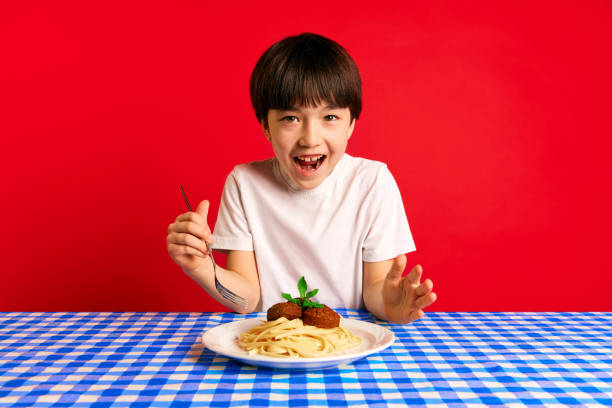 istock Young boy feeling excited, sitting at table with blue checkered tablecloth and plate of spaghetti and meatballs against red background 2153384056