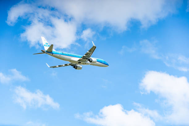 KLM Airlines plane, displaying a blue and white livery, is captured mid-flight against a backdrop of a clear blue sky with scattered white clouds. The aircraft is seen from the rear and below, showing its engines and tailfin as it ascends. The image highlights the plane's upward trajectory and the serene sky.